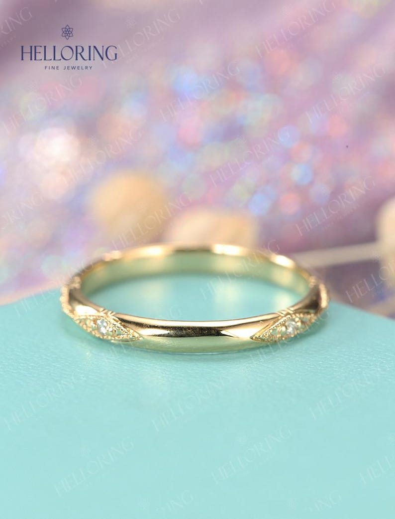 May include: A thin gold wedding band with a delicate diamond pattern. The band is on a light blue background.