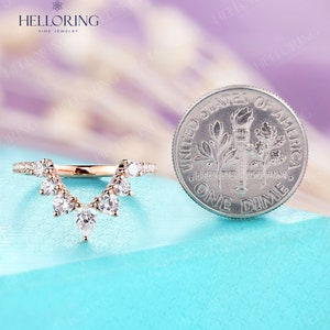 May include: A rose gold wedding band with a V-shaped design featuring a row of small diamonds on each side and a larger diamond at the point of the V. The band is shown next to a US dime for size comparison.
