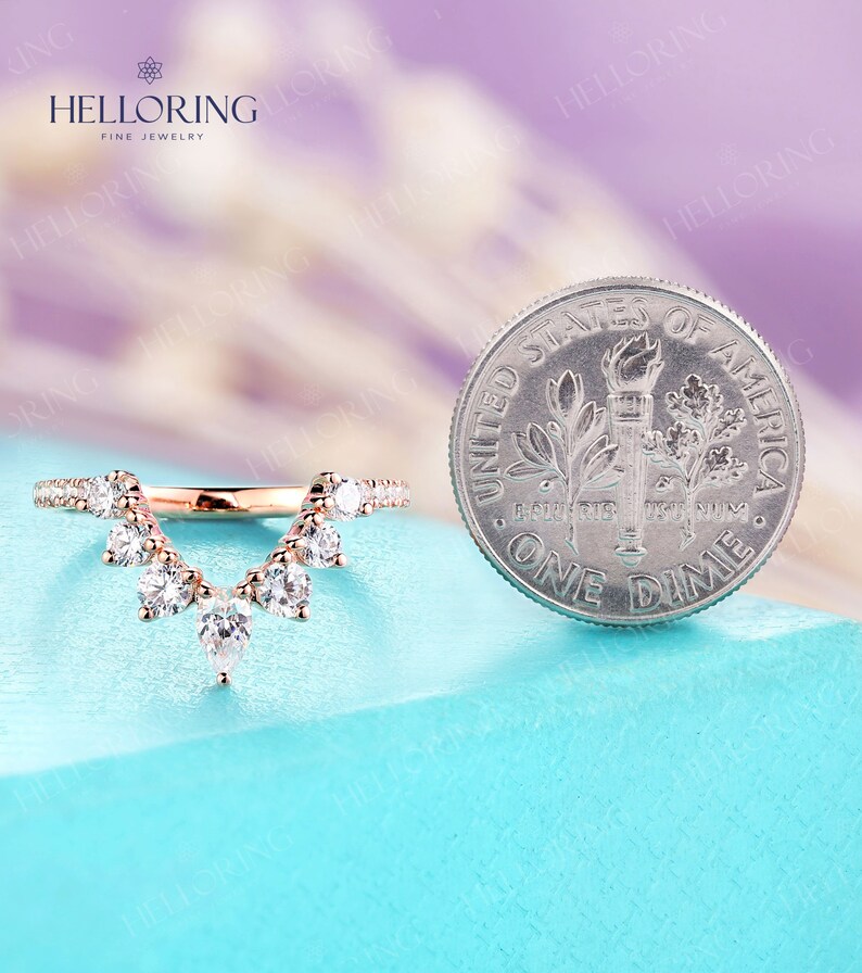 May include: A rose gold wedding band with a V-shaped design featuring a row of small diamonds on each side and a larger diamond at the point of the V. The band is shown next to a US dime for size comparison.