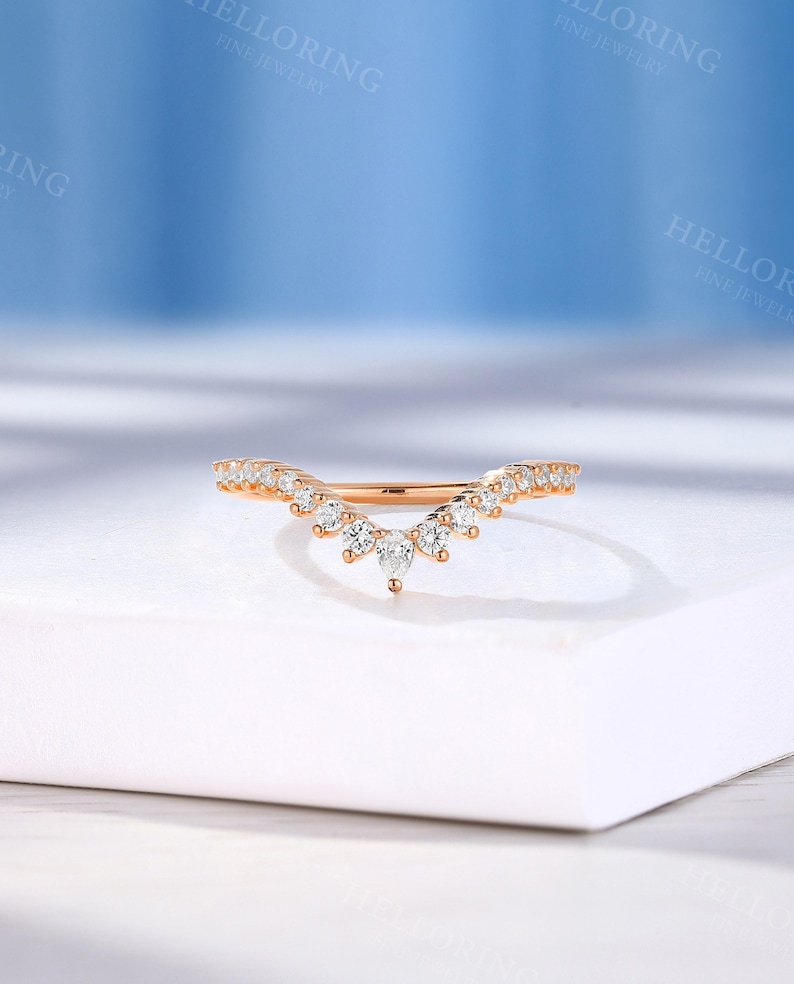 May include: A delicate rose gold wedding band with a curved, v-shaped design. The band is adorned with small, sparkling diamonds.