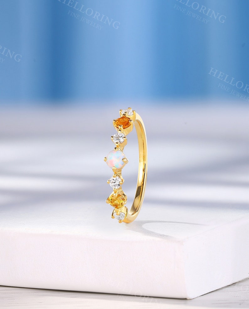 May include: A gold band ring with a row of gemstones. The ring features a white opal, two yellow gemstones, and small clear gemstones.