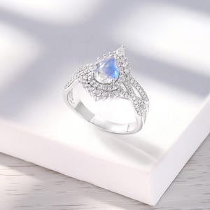 Peut inclure: Une bague en argent avec une pierre de lune en forme de poire entour&eacute;e d'un halo de petites pierres. La bague a un design d&eacute;licat et complexe.