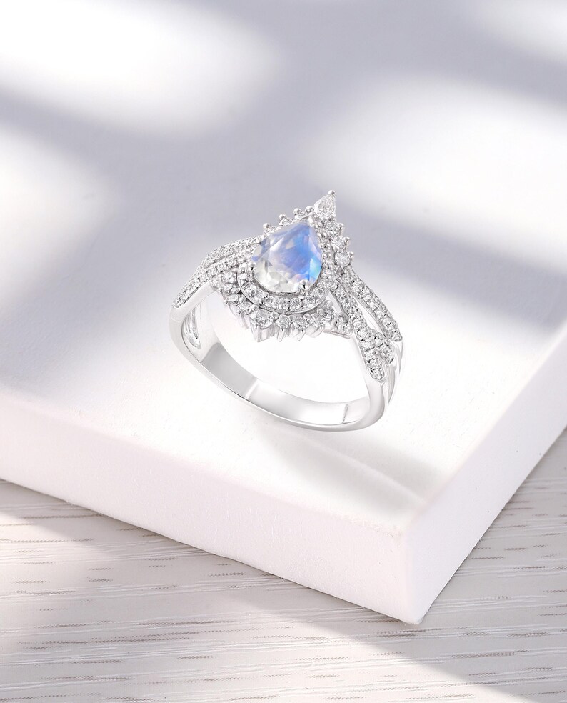 Peut inclure: Une bague en argent avec une pierre de lune en forme de poire entour&eacute;e d'un halo de petites pierres. La bague a un design d&eacute;licat et complexe.