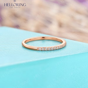 May include: Rose gold band ring with a row of small, clear gemstones set along the top. The ring is displayed on a light blue surface, with the brand name "HELLORING FINE JEWELRY" visible in the background.