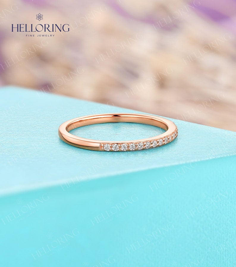 May include: Rose gold band ring with a row of small, clear gemstones set along the top. The ring is displayed on a light blue surface, with the brand name "HELLORING FINE JEWELRY" visible in the background.