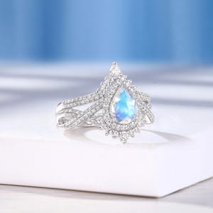Peut inclure: Une bague de fian&ccedil;ailles en argent avec une pierre de lune en forme de poire et un halo de petits diamants. La bague est pos&eacute;e sur une surface blanche.