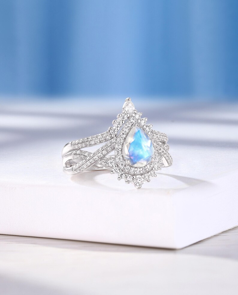 Peut inclure: Une bague de fian&ccedil;ailles en argent avec une pierre de lune en forme de poire et un halo de petits diamants. La bague est pos&eacute;e sur une surface blanche.