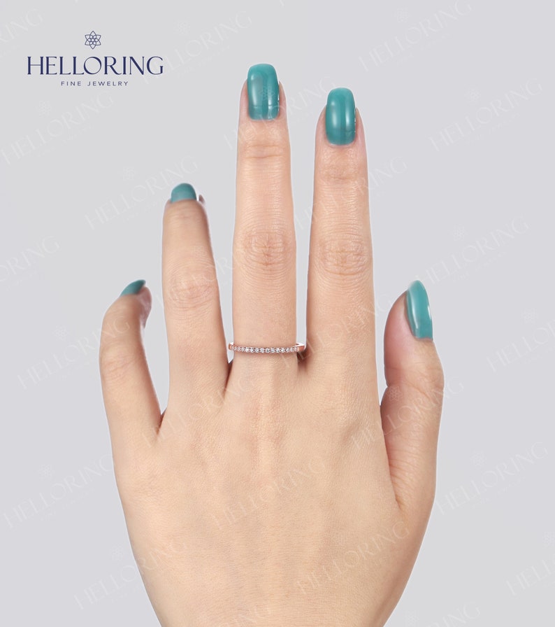 May include: A rose gold band ring with small, clear gemstones set across the top. The ring is displayed on a hand with turquoise nail polish. The ring is from the brand HELLORING FINE JEWELRY.