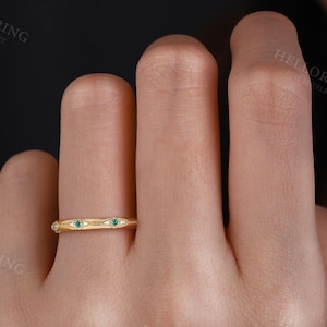 May include: A gold band ring featuring a series of repeating marquise-shaped engravings, each centered with a small, round, green gemstone. The metal surface displays a polished finish with delicate beaded detailing along the edges of the motifs.