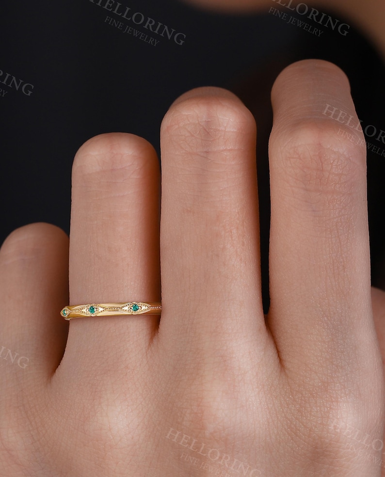 May include: A gold band ring featuring a series of repeating marquise-shaped engravings, each centered with a small, round, green gemstone. The metal surface displays a polished finish with delicate beaded detailing along the edges of the motifs.