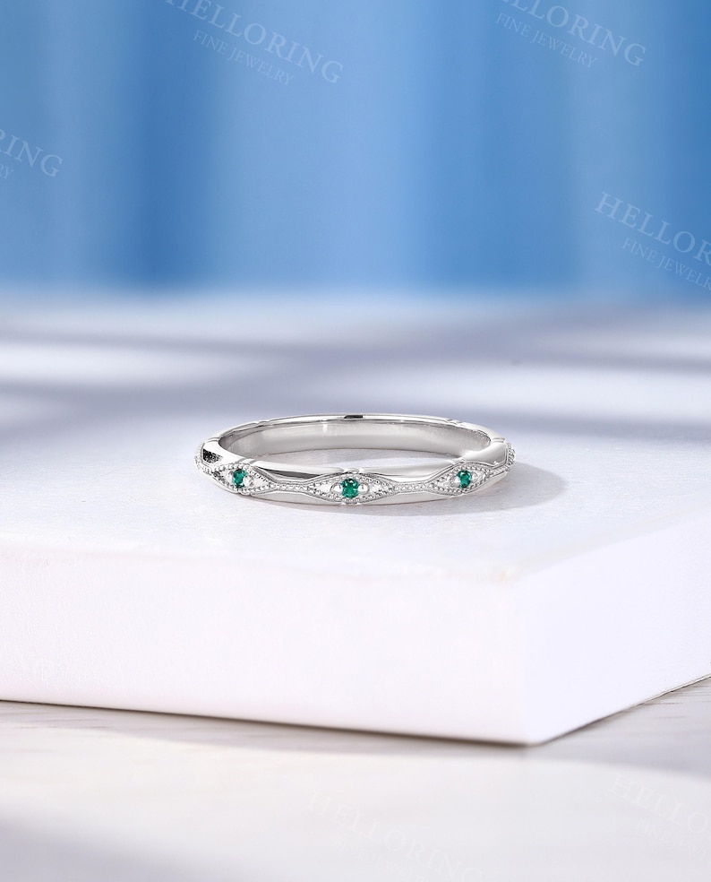 May include: A silver-toned metal band ring featuring a series of marquise-shaped engravings, each set with a small, round, vibrant green gemstone. The band displays intricate milgrain detailing along the edges of the marquise motifs.