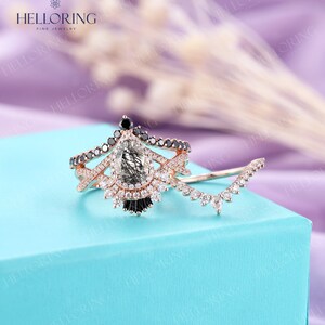 May include: A pear-shaped engagement ring with a black diamond halo and a pave band. The ring is set in rose gold and features a delicate, curved band with smaller diamonds.