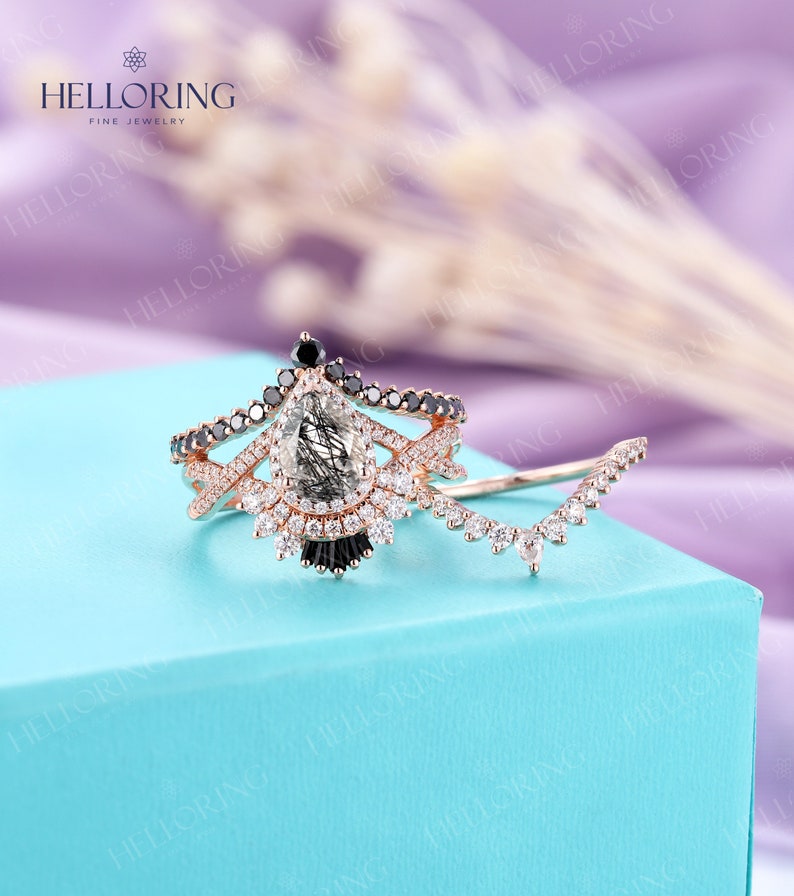 May include: A pear-shaped engagement ring with a black diamond halo and a pave band. The ring is set in rose gold and features a delicate, curved band with smaller diamonds.