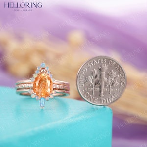 May include: A pear-shaped sunstone engagement ring with a halo of white opal stones and two opal accents on the band. The ring is set in rose gold and is shown next to a US dime for size comparison.