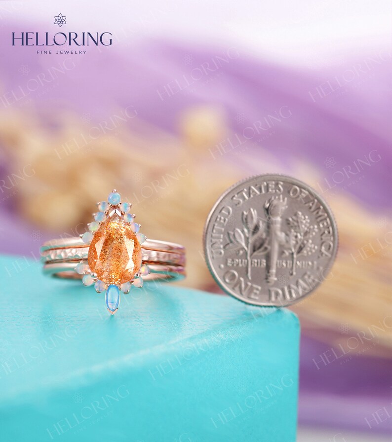 May include: A pear-shaped sunstone engagement ring with a halo of white opal stones and two opal accents on the band. The ring is set in rose gold and is shown next to a US dime for size comparison.