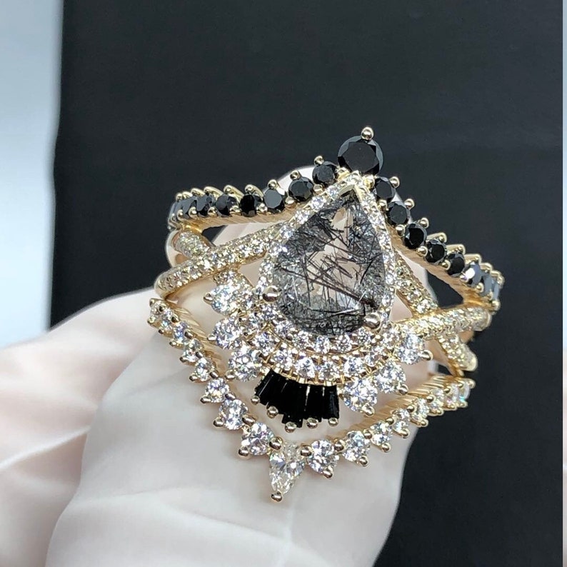 May include: A gold ring set with a pear-shaped clear quartz stone with black inclusions. The ring is surrounded by a halo of smaller clear stones and black stones. The ring is part of a set of three rings.