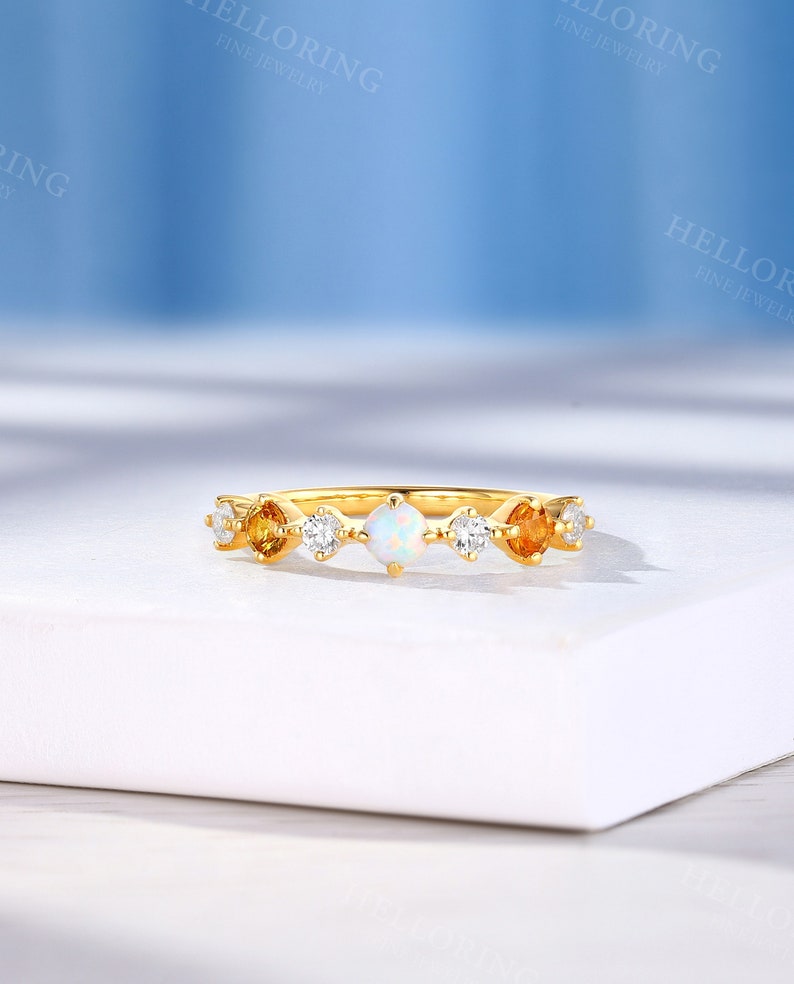 May include: A gold band ring with a white opal and small yellow gemstones. The ring is on a white surface.