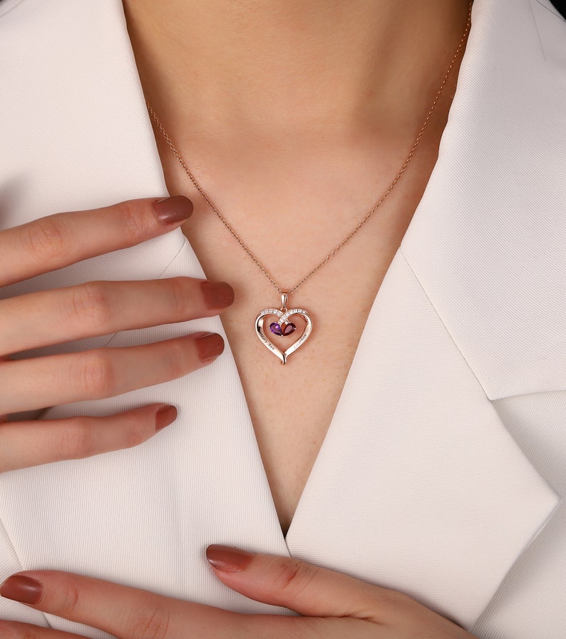 May include: Rose gold heart pendant necklace with a fine chain. The pendant has a heart-shaped design with a smaller heart inside, set with purple gemstones and small white stones. The necklace is worn against a white blazer.