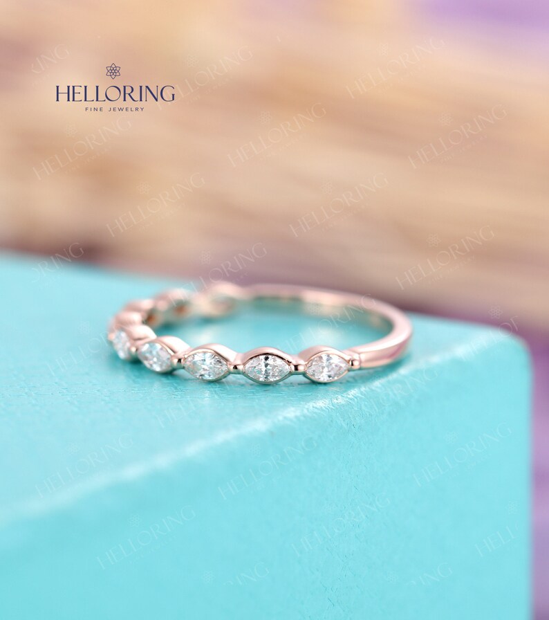 May include: A delicate rose gold band with a row of marquise-shaped diamonds. The band is set on a light blue background.