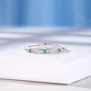 May include: A silver-toned metal band ring featuring a series of small, round, vibrant green gemstones set within delicate, diamond-shaped milgrain detailing. The ring has a polished finish and a slim, uniform profile, showcasing a repeating pattern of stones.