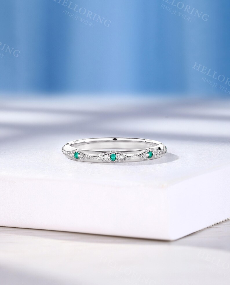 May include: A silver-toned metal band ring featuring a series of small, round, vibrant green gemstones set within delicate, diamond-shaped milgrain detailing. The ring has a polished finish and a slim, uniform profile, showcasing a repeating pattern of stones.