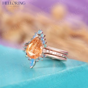 May include: A rose gold ring set with a pear-shaped sunstone and a halo of smaller opal stones. The ring is on a turquoise surface.