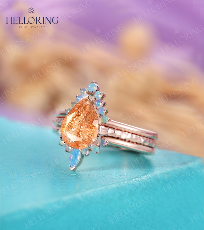 May include: A rose gold ring set with a pear-shaped sunstone and a halo of smaller opal stones. The ring is on a turquoise surface.