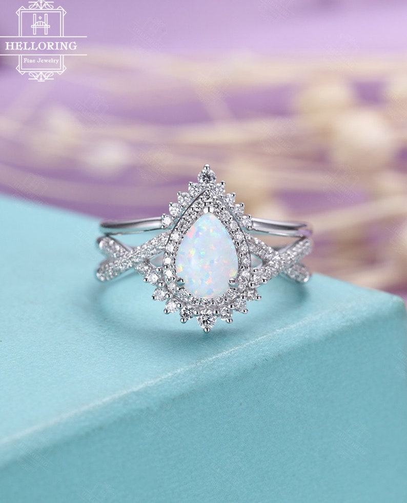 May include: A pear-shaped opal engagement ring with a halo of smaller diamonds. The ring is set in silver and has a delicate, intricate design. The ring is on a light blue background.