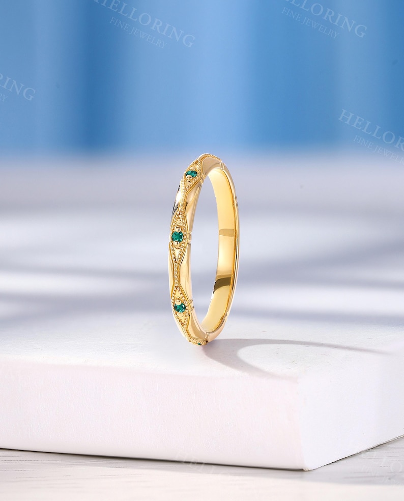 May include: A yellow gold band ring featuring a series of small, round, green gemstones set within decorative, diamond-shaped metal frames. The band has a polished finish and intricate milgrain detailing along the edges of the settings.