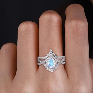Peut inclure: Une bague de fian&ccedil;ailles en or blanc avec une pierre de lune en forme de poire et un halo de diamants plus petits. La bague est sertie d'un anneau en or blanc avec un design d&eacute;licat et torsad&eacute;.