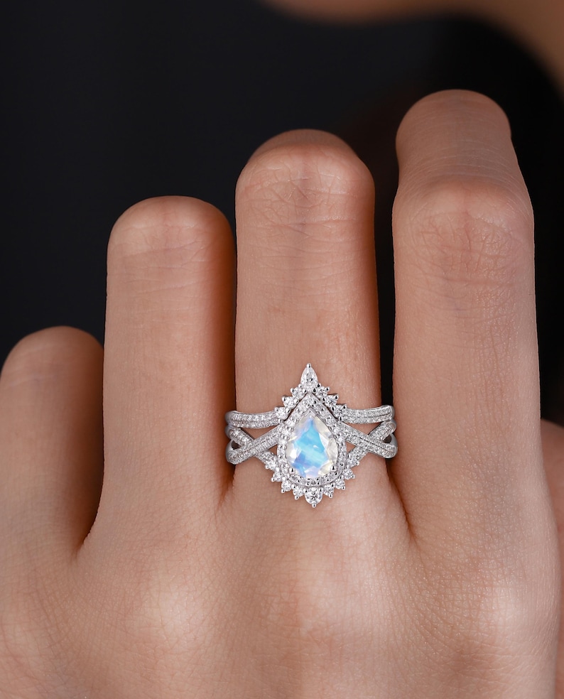Peut inclure: Une bague de fian&ccedil;ailles en or blanc avec une pierre de lune en forme de poire et un halo de diamants plus petits. La bague est sertie d'un anneau en or blanc avec un design d&eacute;licat et torsad&eacute;.