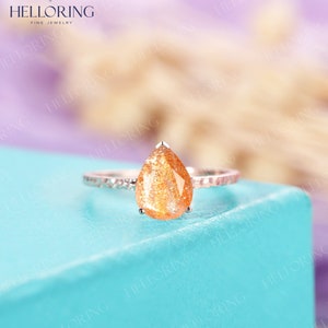 May include: A pear-shaped sunstone engagement ring set in a silver band with a textured finish. The ring is on a light blue surface.