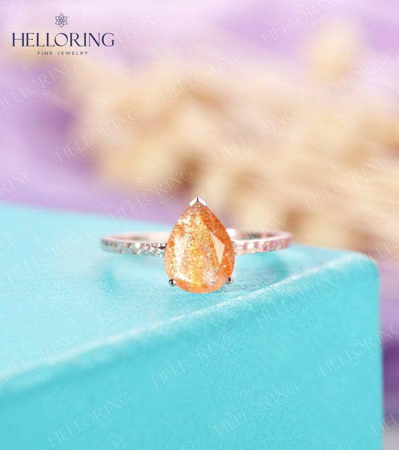 May include: A pear-shaped sunstone engagement ring set in a silver band with a textured finish. The ring is on a light blue surface.