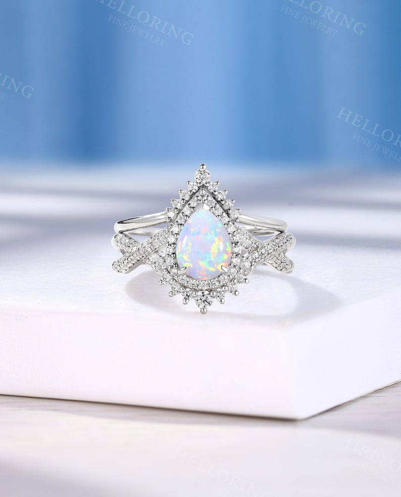 May include: A silver engagement ring set with a pear-shaped opal center stone, surrounded by a halo of small, clear gemstones. The band features additional small gemstones. The ring is set against a white background.