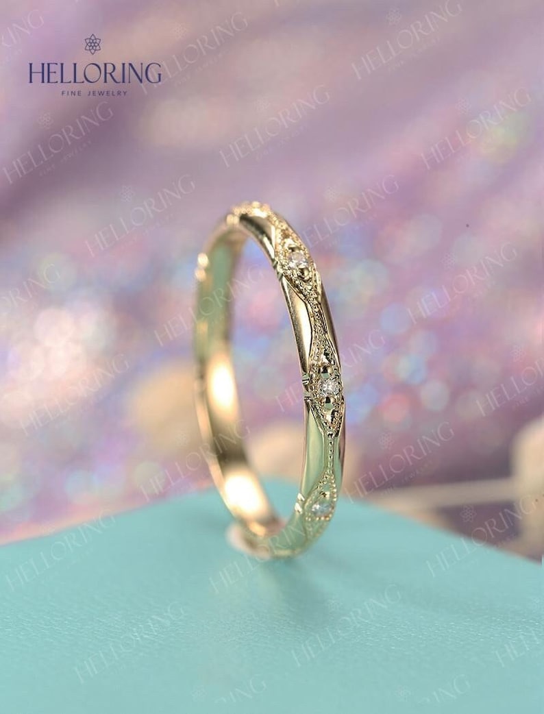 May include: A delicate gold wedding band with a decorative design and small diamonds. The band is on a light blue background.