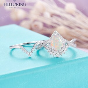 May include: A pear-shaped opal engagement ring with a halo of small diamonds. The ring is set in white gold and has a delicate, twisted band. The ring is paired with a matching wedding band that features a V-shaped design with small diamonds.