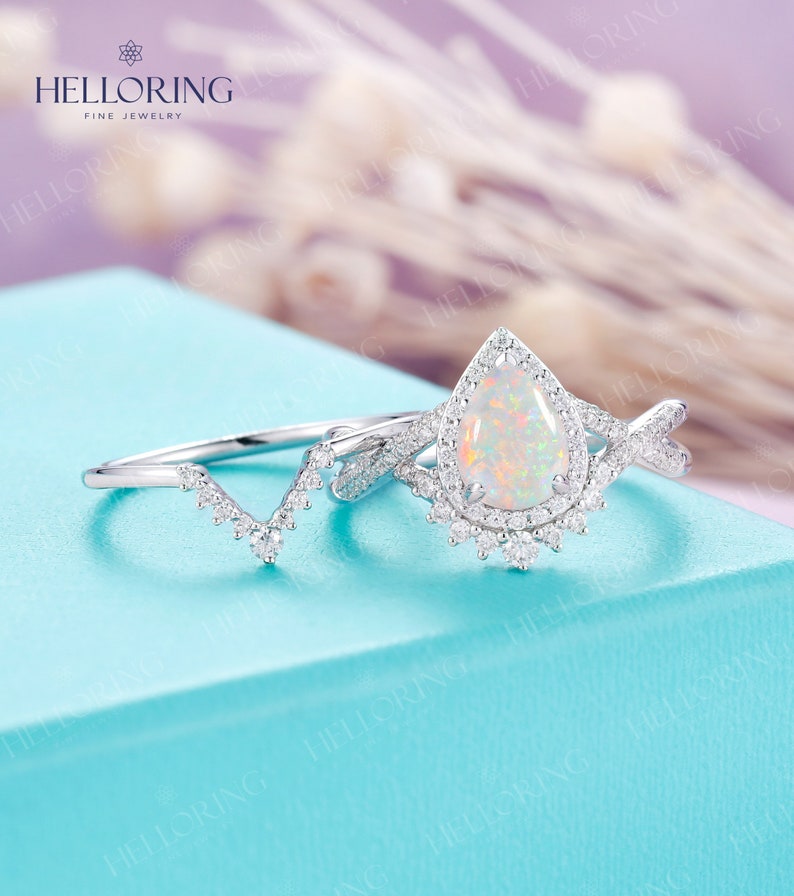 May include: A pear-shaped opal engagement ring with a halo of small diamonds. The ring is set in white gold and has a delicate, twisted band. The ring is paired with a matching wedding band that features a V-shaped design with small diamonds.