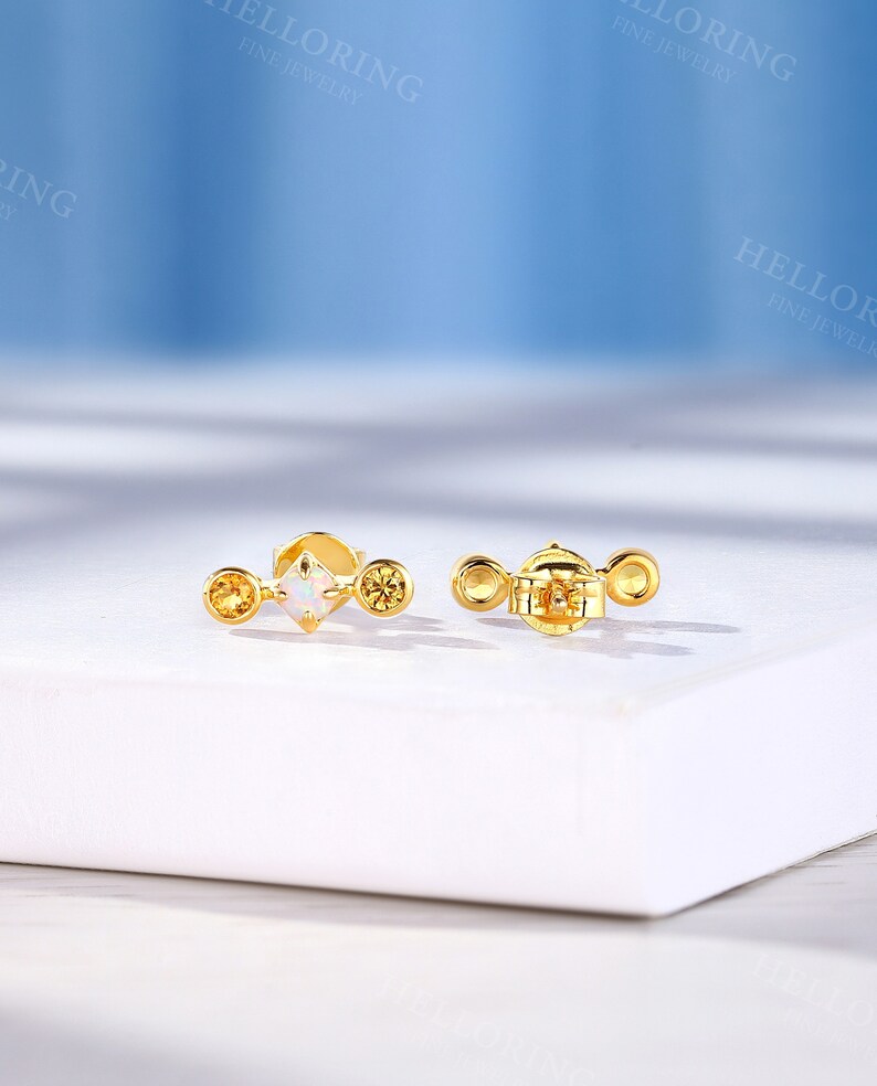 May include: A pair of gold earrings with a small white opal gemstone and two smaller yellow gemstones. The earrings are on a white surface.