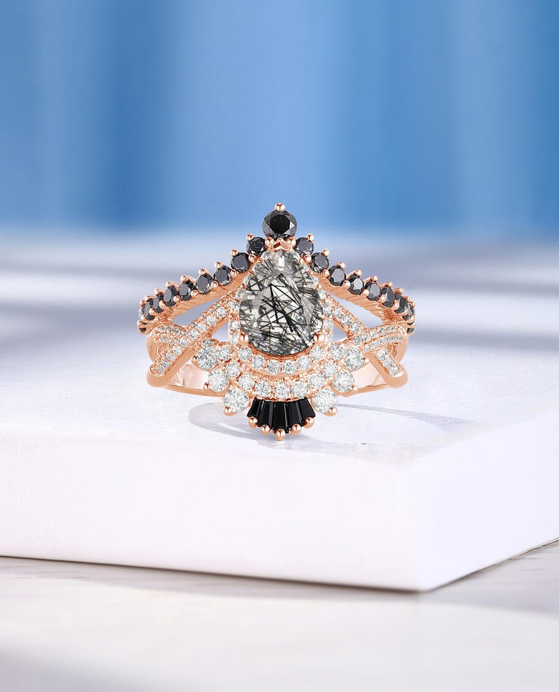 May include: A rose gold ring with a pear-shaped clear stone surrounded by smaller clear stones. The ring has a band with black stones and a design that resembles a crown.