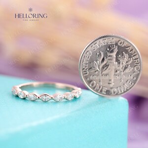 May include: A delicate, thin wedding band with a row of marquise-shaped diamonds set in a scalloped edge. The band is shown next to a US dime for size comparison.