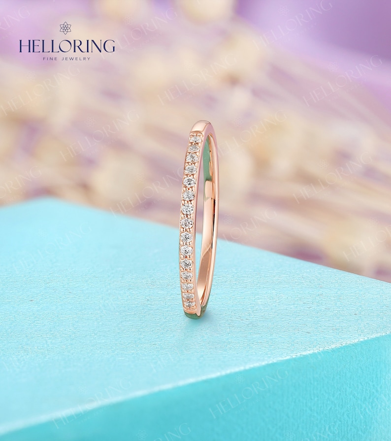 May include: A rose gold band ring with small, clear gemstones set along the top. The ring is displayed on a light blue surface. The brand name "HELLORING FINE JEWELRY" is visible in the upper left corner.