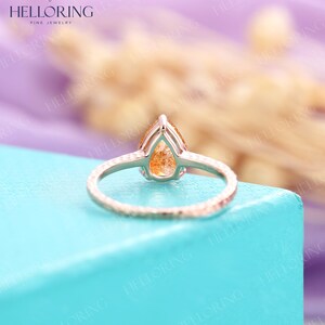 May include: A pear-shaped, orange gemstone ring set in a silver band with a textured finish. The ring is on a light blue surface.