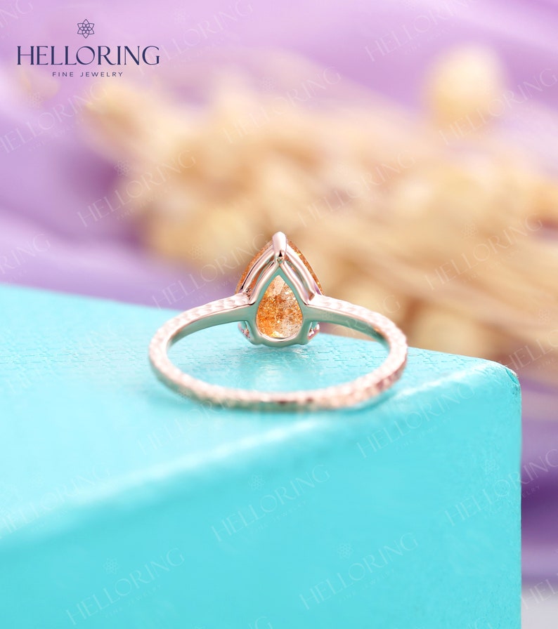 May include: A pear-shaped, orange gemstone ring set in a silver band with a textured finish. The ring is on a light blue surface.