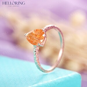 May include: A pear-shaped orange sunstone ring with a textured band. The ring is set in a silver setting and is on a light blue surface.