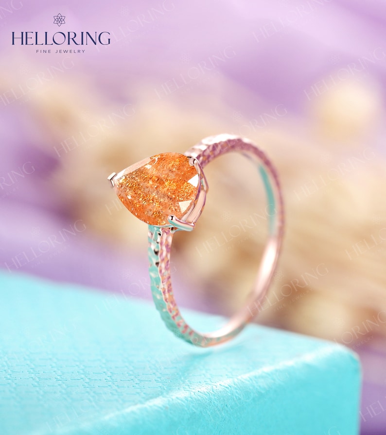 May include: A pear-shaped orange sunstone ring with a textured band. The ring is set in a silver setting and is on a light blue surface.