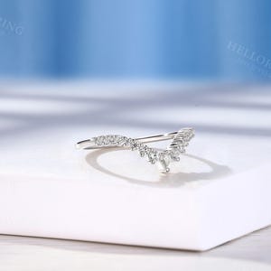 May include: A delicate silver ring with a V-shaped design featuring a row of small sparkling diamonds. The ring is set on a white surface.