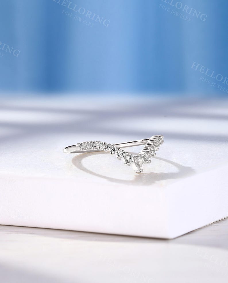May include: A delicate silver ring with a V-shaped design featuring a row of small sparkling diamonds. The ring is set on a white surface.