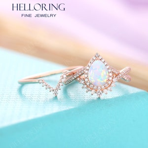 May include: A rose gold engagement ring set with a pear-shaped opal center stone and a delicate band. The ring is surrounded by a halo of smaller diamonds. The set also includes a matching wedding band with a delicate design.