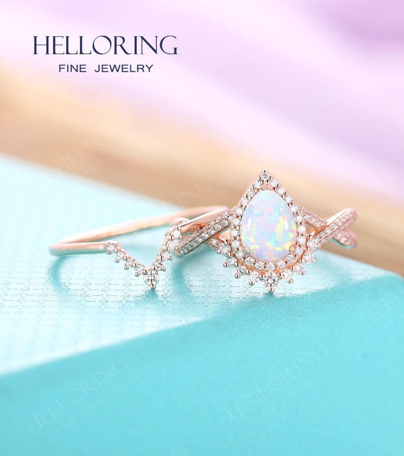 May include: A rose gold engagement ring set with a pear-shaped opal center stone and a delicate band. The ring is surrounded by a halo of smaller diamonds. The set also includes a matching wedding band with a delicate design.