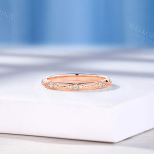 May include: A rose gold band ring featuring a series of small, round, clear stones set within a delicate, engraved metal pattern. The ring has a polished finish and a slim profile, showcasing intricate detailing along the outer surface.